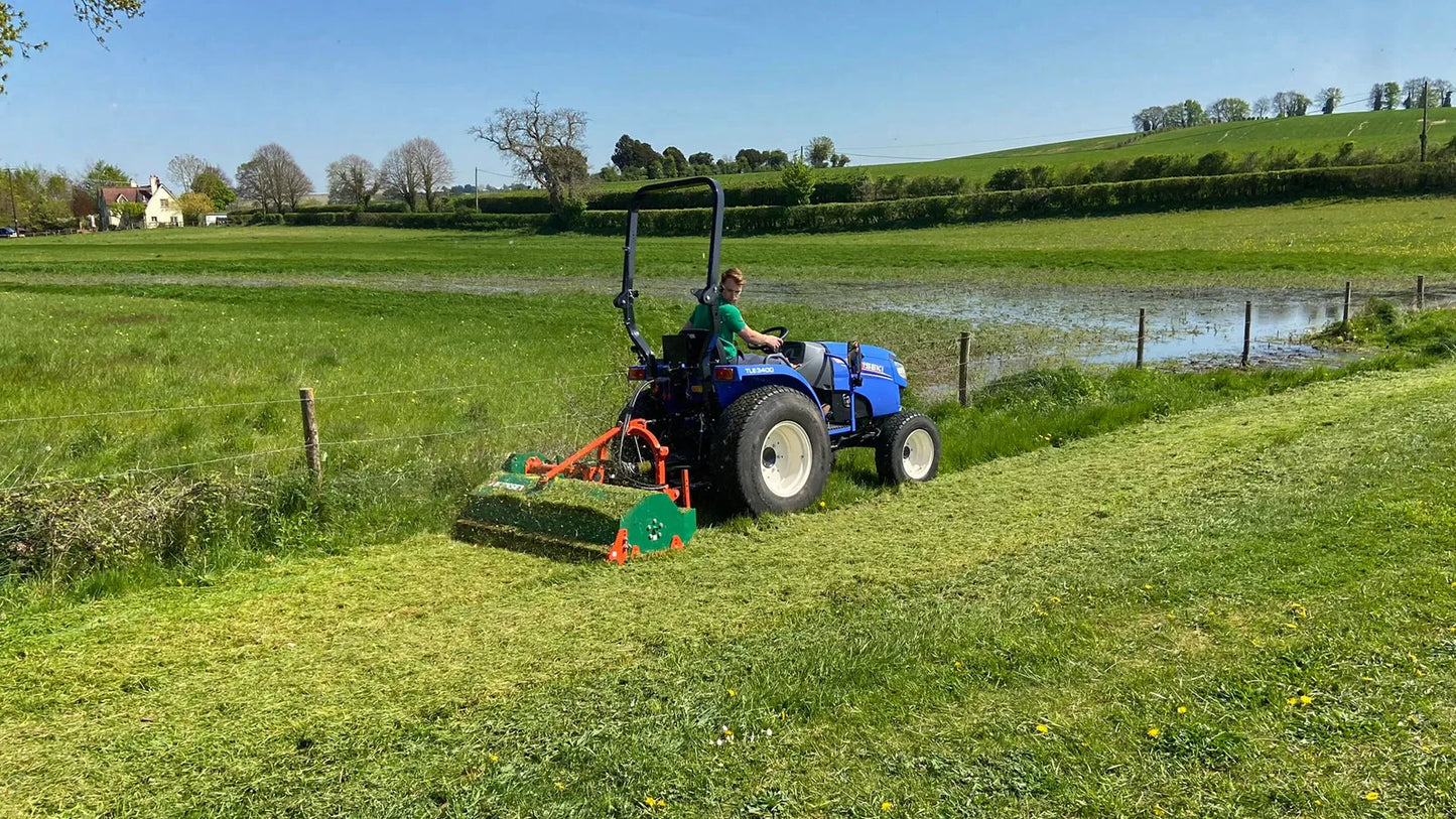 Tractor mowing a field with a pond and trees in the background