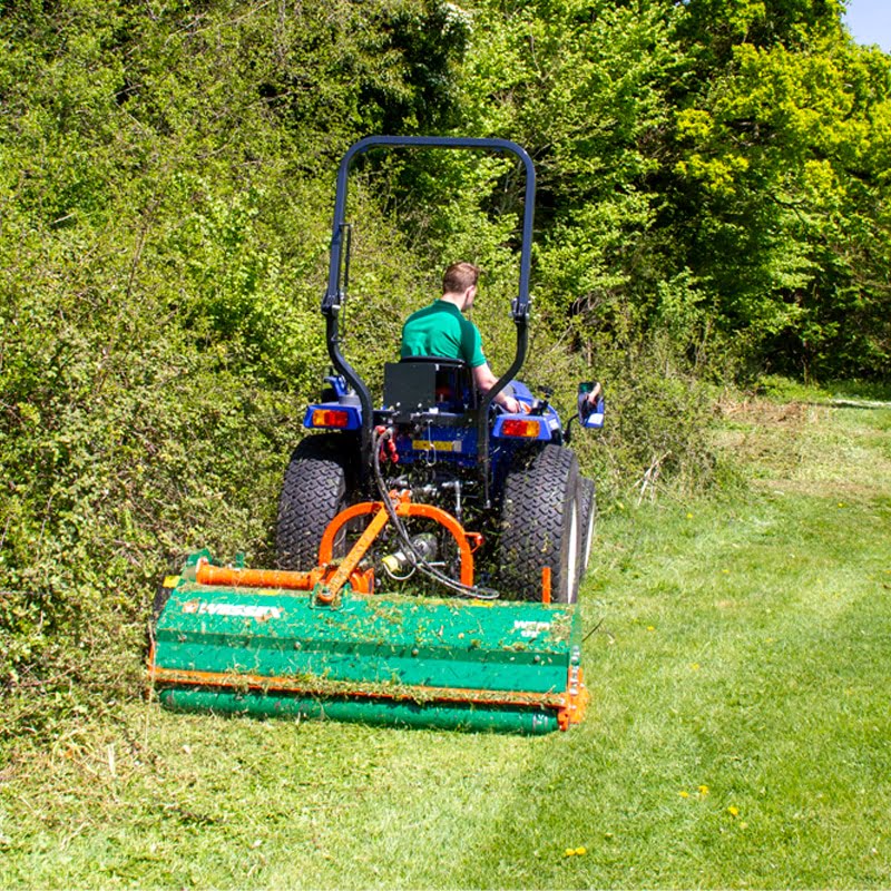 Person operating a tractor with a large green Wessex Flail Mower attachment in a grassy area with trees.