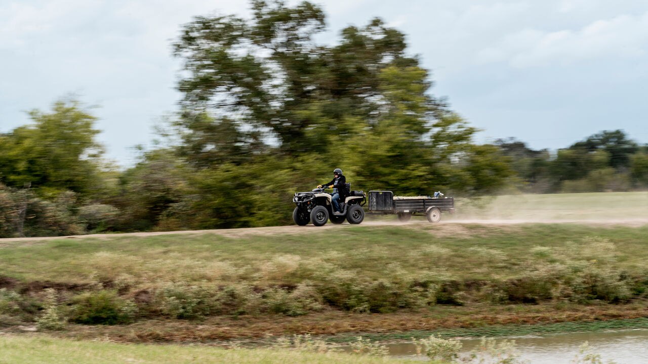 Person riding a Can-Am ATV with a trailer in a grassy field with trees in the background