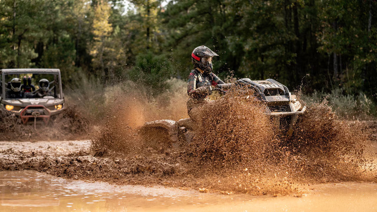 Two Can-Am off-road vehicles driving through a muddy forest path with trees in the background.