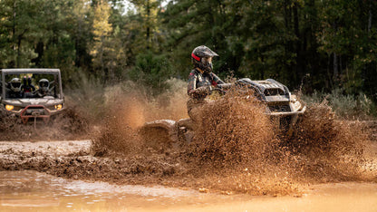 Two Can-Am off-road vehicles driving through a muddy forest path with trees in the background.
