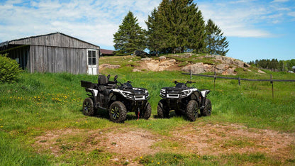 Two Can-Am Outlander Electric all-terrain vehicles parked on a grassy field with a wooden shed and trees in the background.