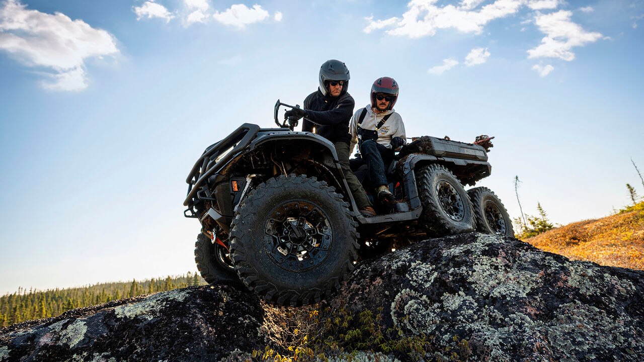 Two people on a Can-Am Outlander 6x6 ATV in a natural landscape with a clear sky.