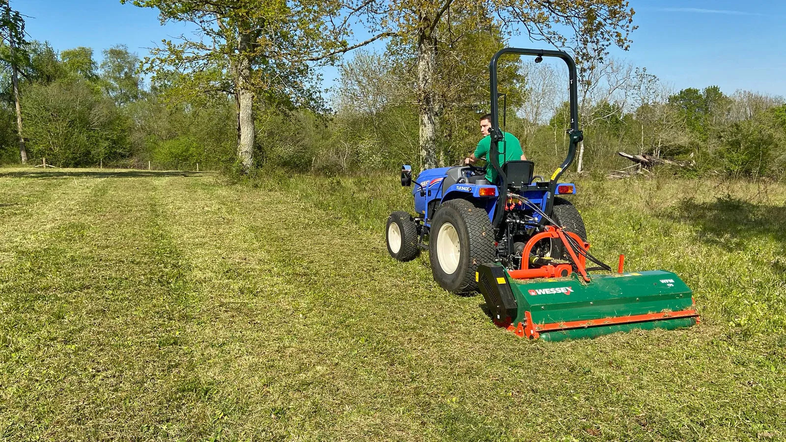 Tractor with a Wessex Flail Mower attachment mowing grass in a field with trees in the background